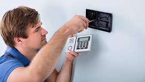 Technician installing a burglar alarm control panel on an interior wall, connecting security sensors for intrusion detection.